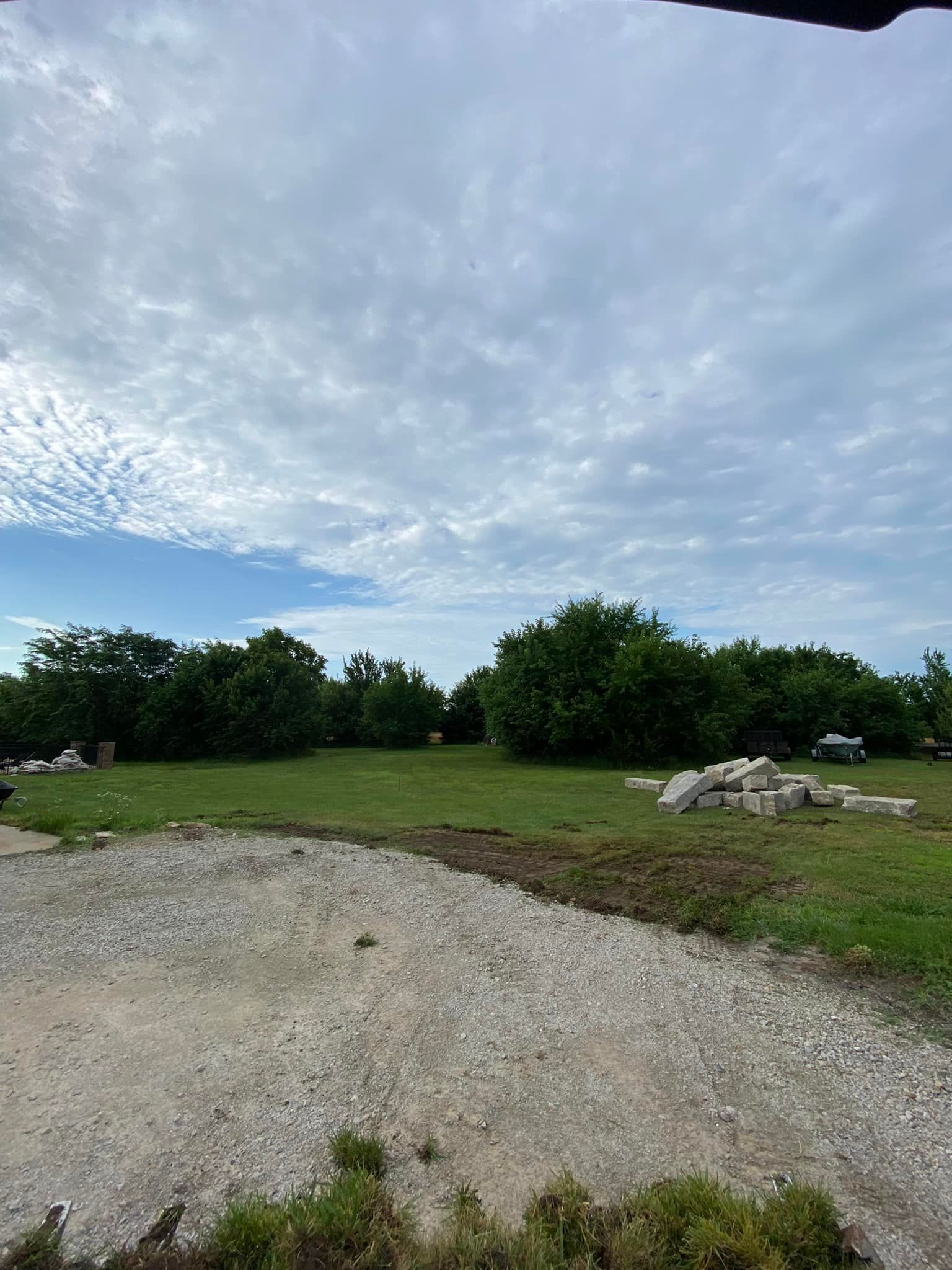 Gravel path beside a grassy field with trees and rocks under a partly cloudy sky