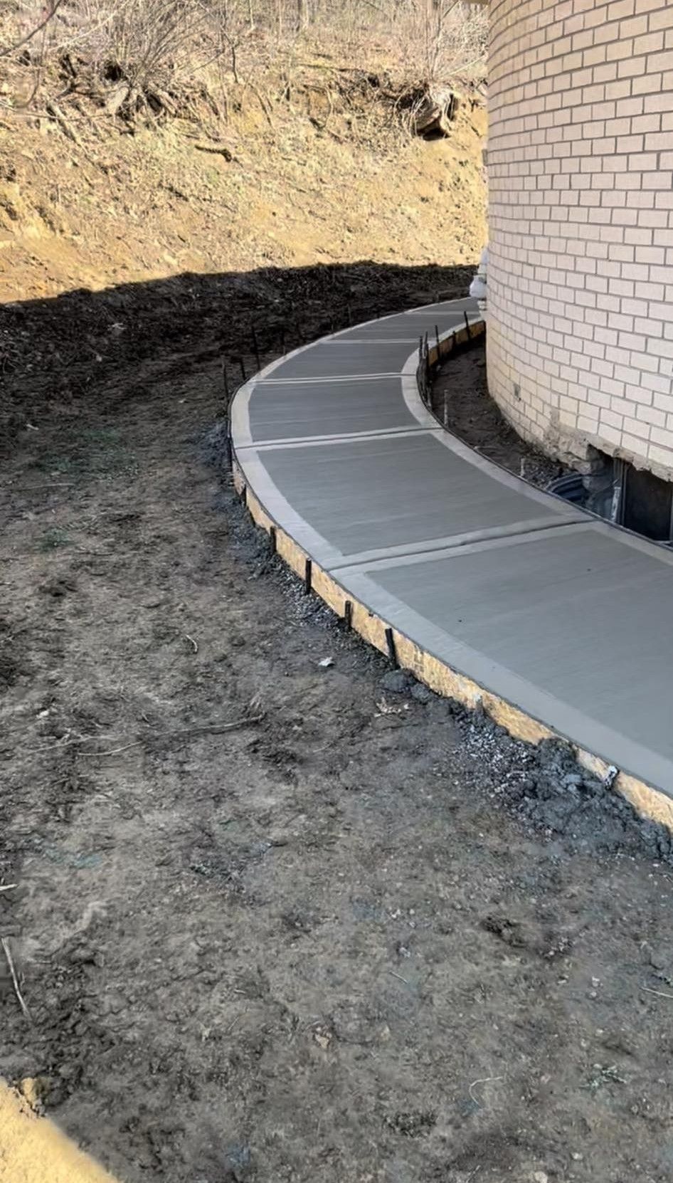 Curved concrete walkway beside a brick building on a dirt hillside, with a dark retaining edge.