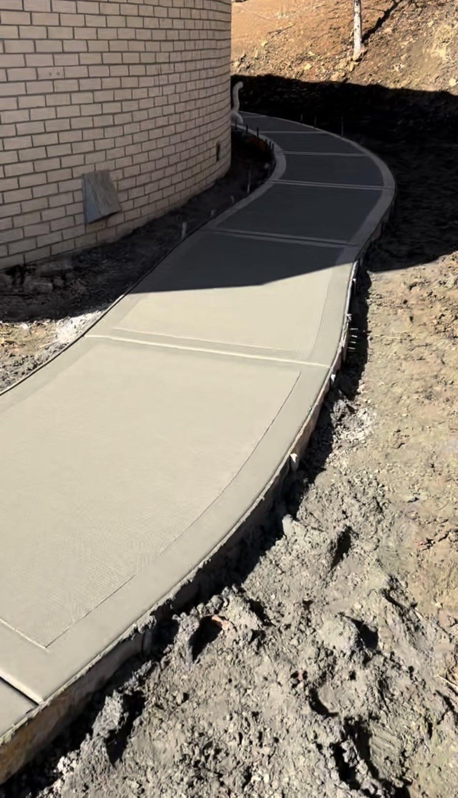 Curved concrete walkway beside a brick wall, bordered by bare dirt and construction debris.