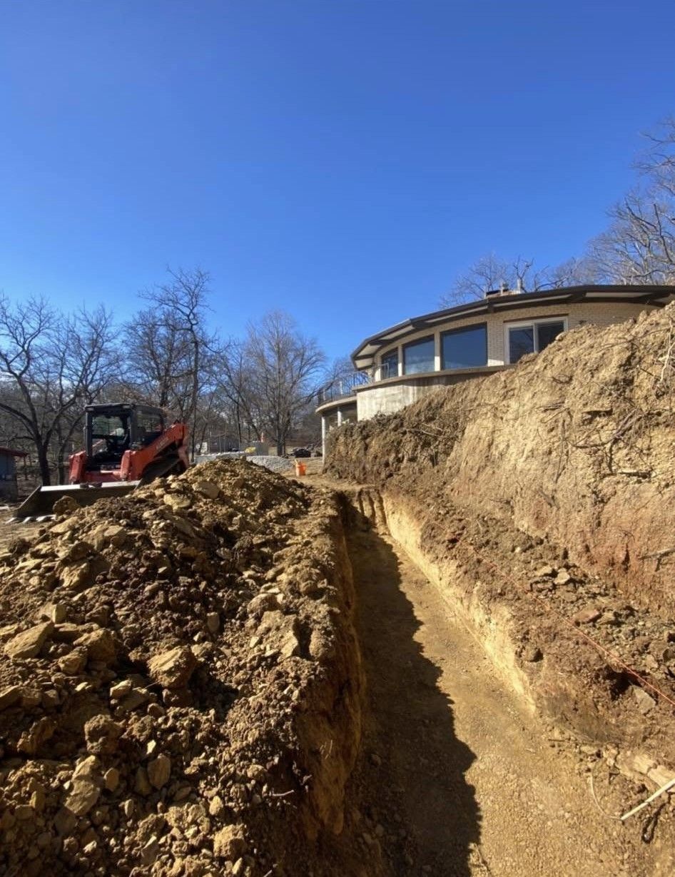 Excavator digging a trench beside a house on a dirt hillside under a blue sky