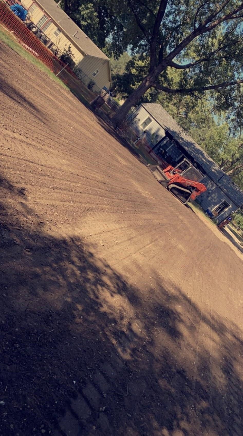 Tilted outdoor scene of a dirt lot with a small orange excavator under trees and buildings in the background