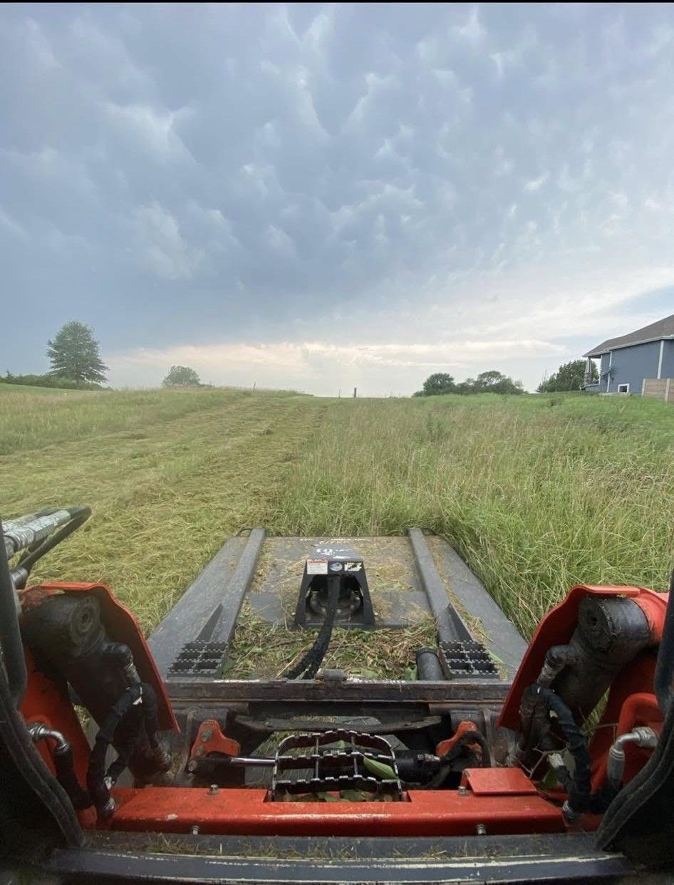 View from a farm machine over a grassy field under cloudy sky, with a barn on the right.