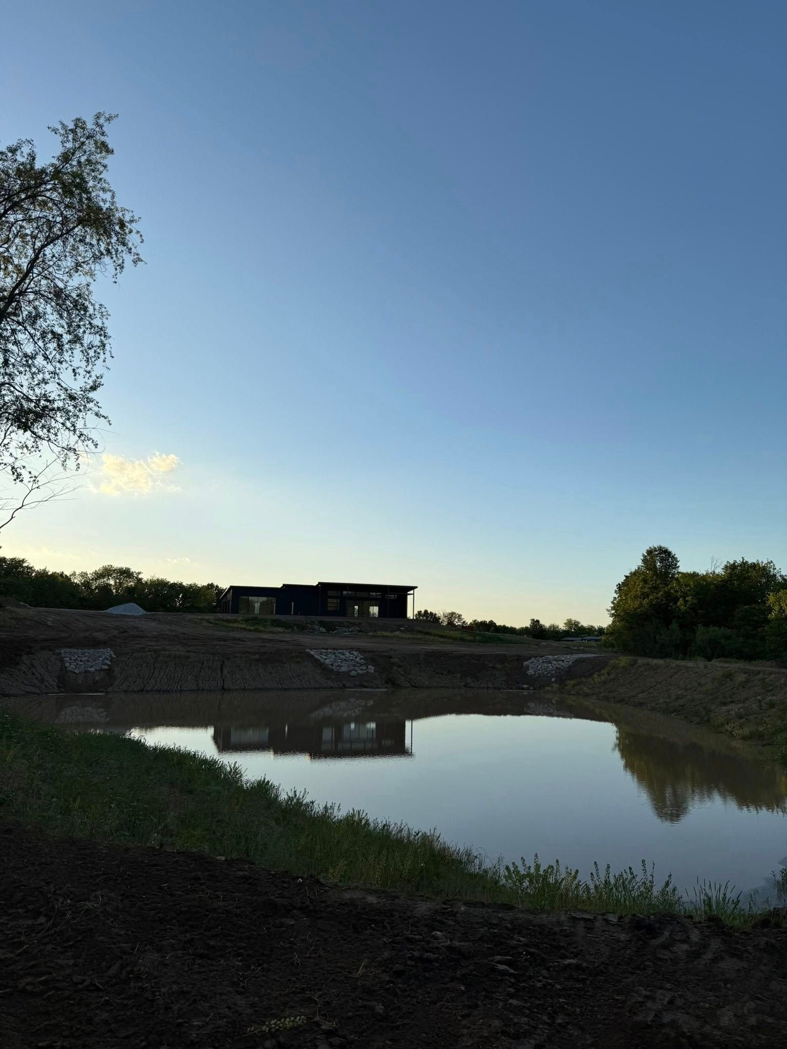 Calm pond at dusk reflecting a dark building and trees under a clear blue sky.