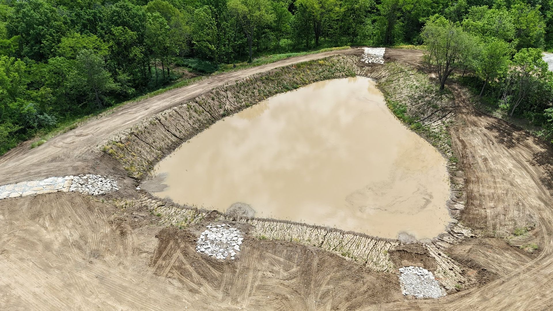 Small muddy pond or excavation pit surrounded by dirt and green trees.