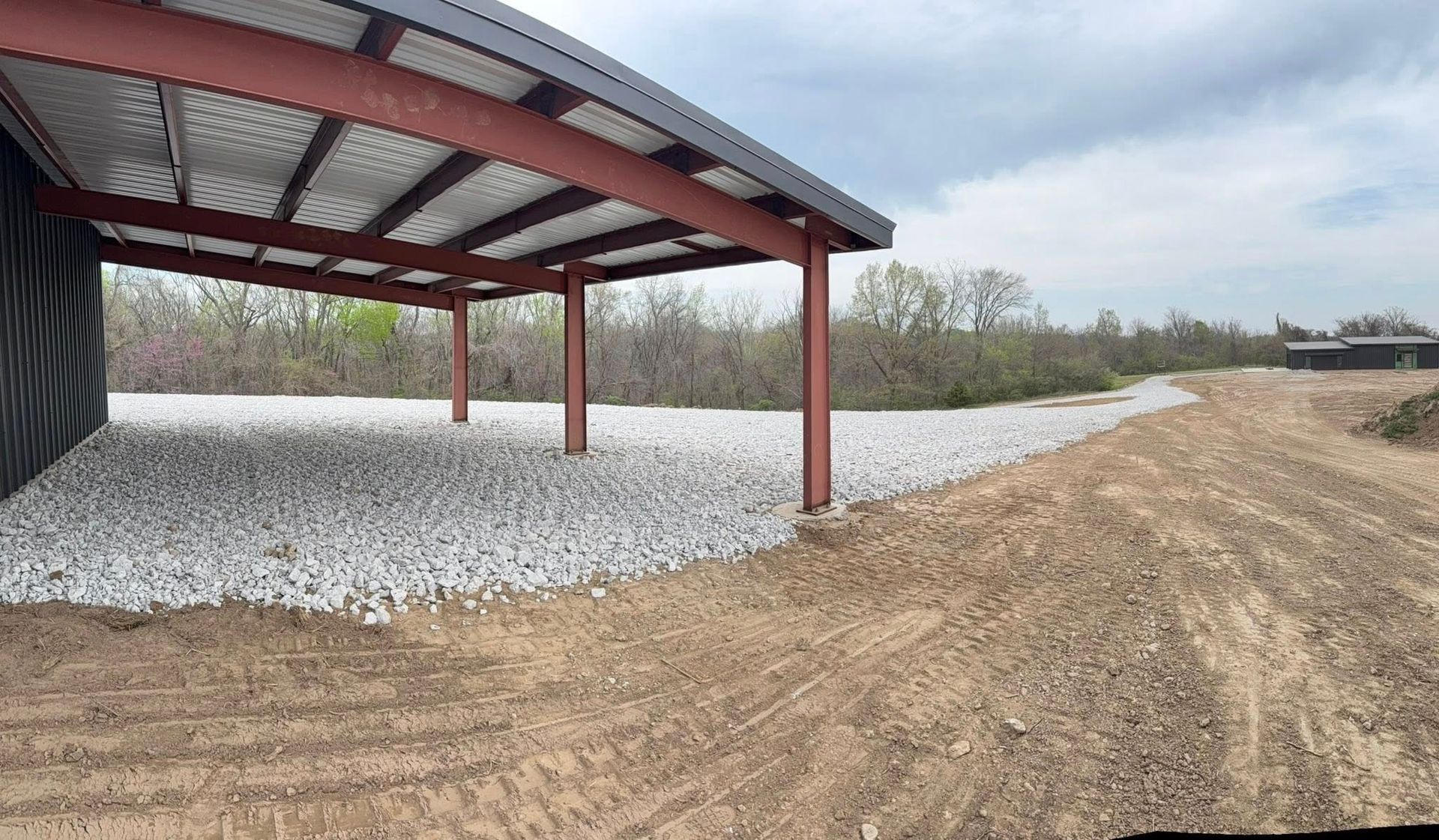 Metal carport on gravel lot beside a dirt road under an overcast sky