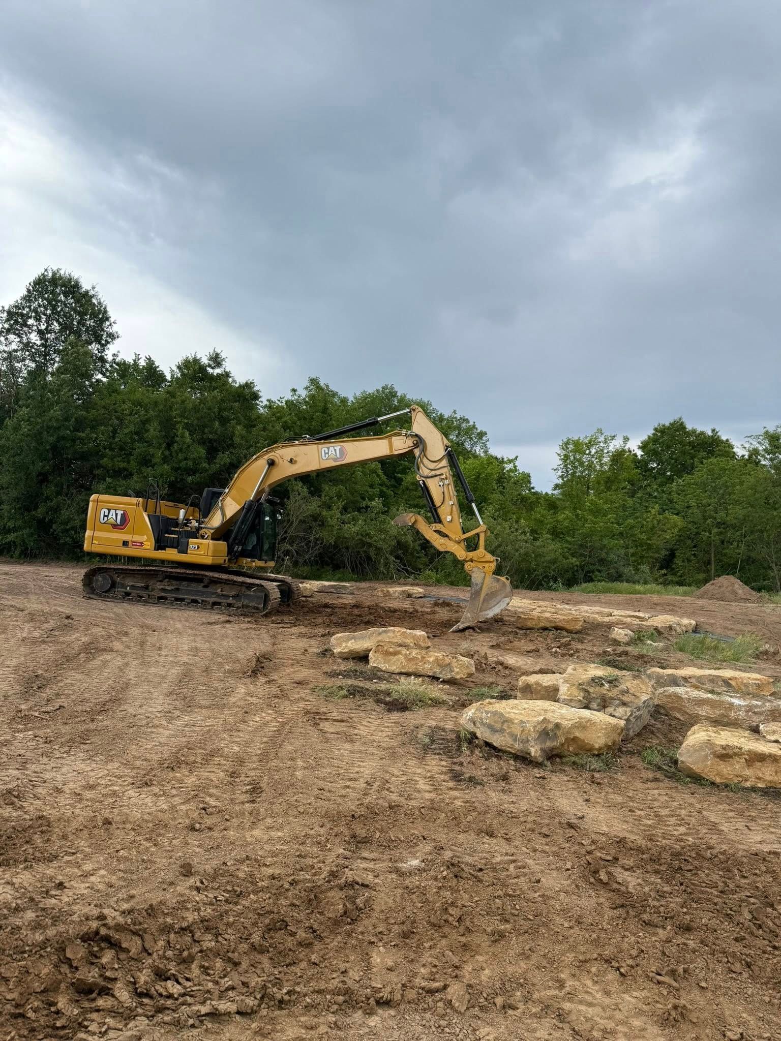 Yellow excavator digging in a muddy field beside a pile of rocks under a cloudy sky