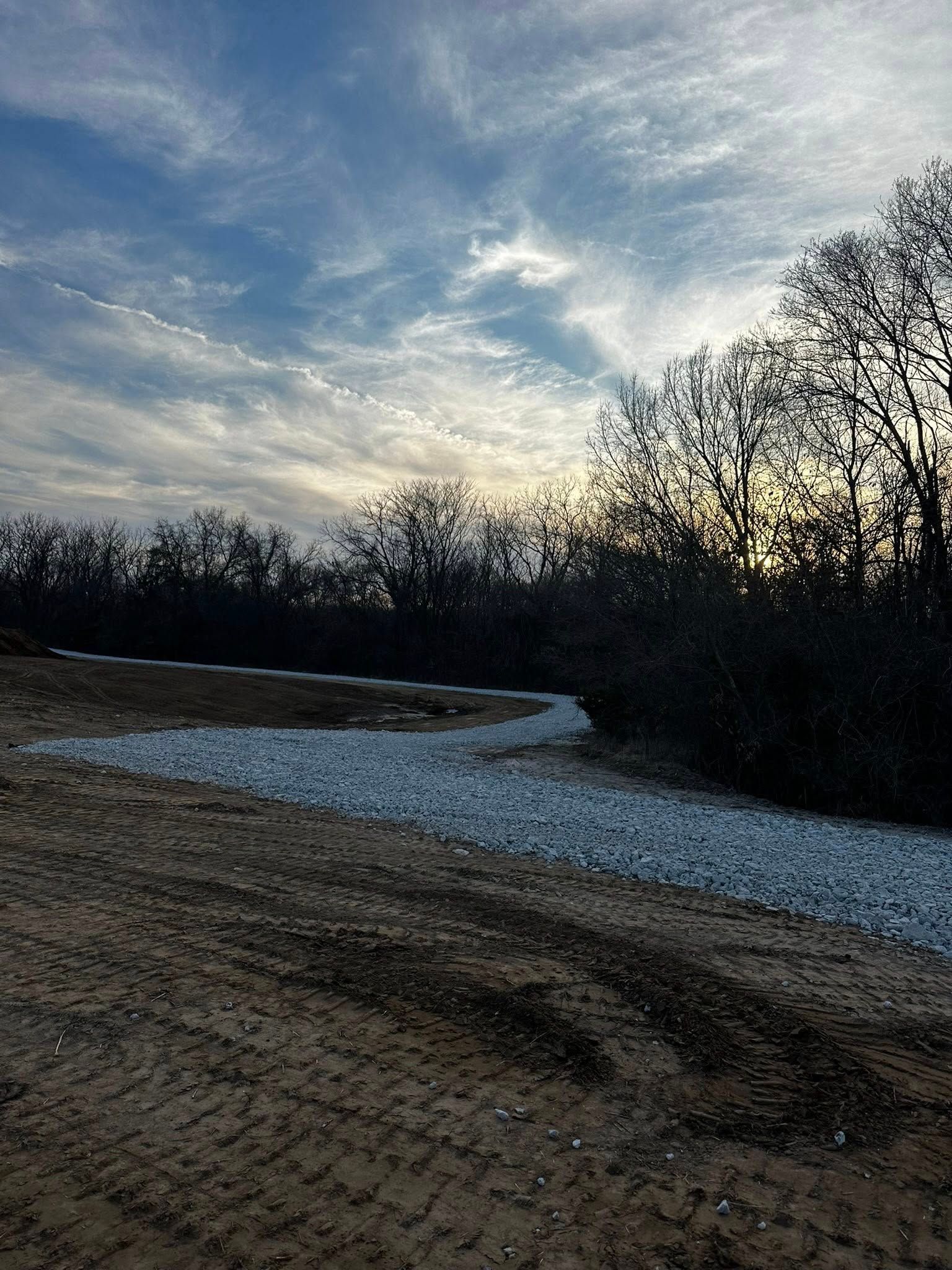 Gravel road curving through bare trees under a cloudy dusk sky