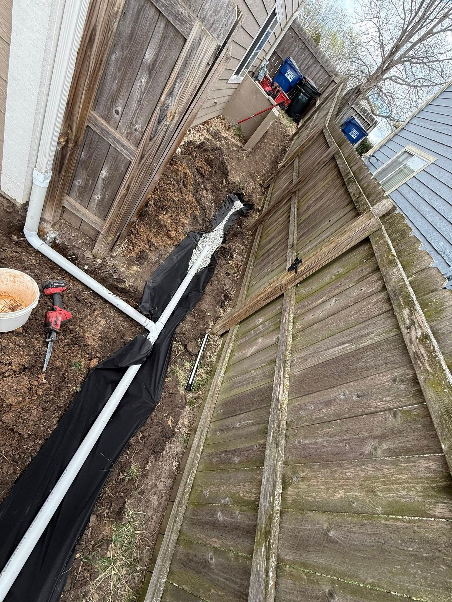 Narrow backyard with wooden fences, gravel ground, and black drainage pipes along a corner.