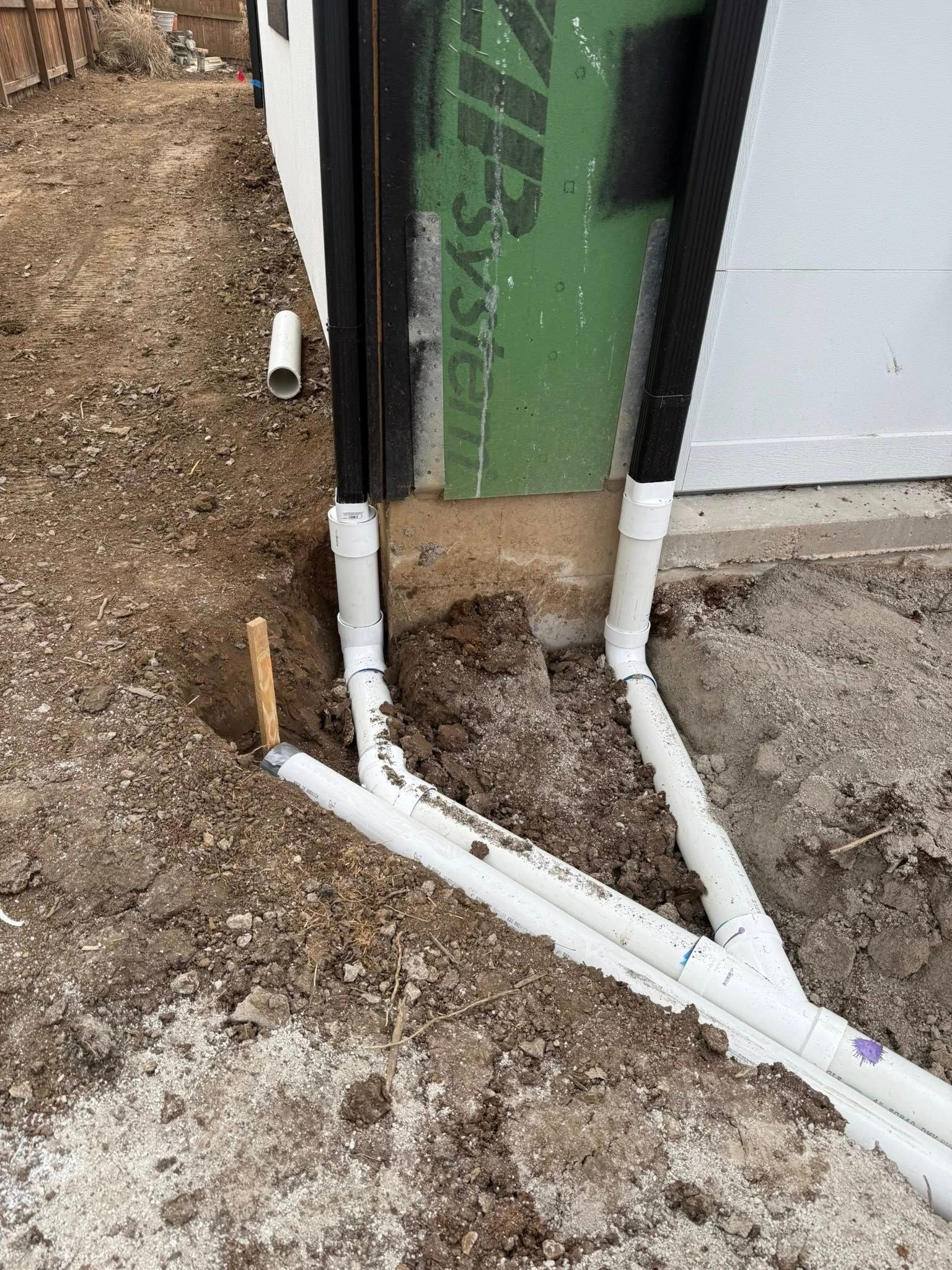 PVC pipes and conduit running along a dirt area beside a green building wall