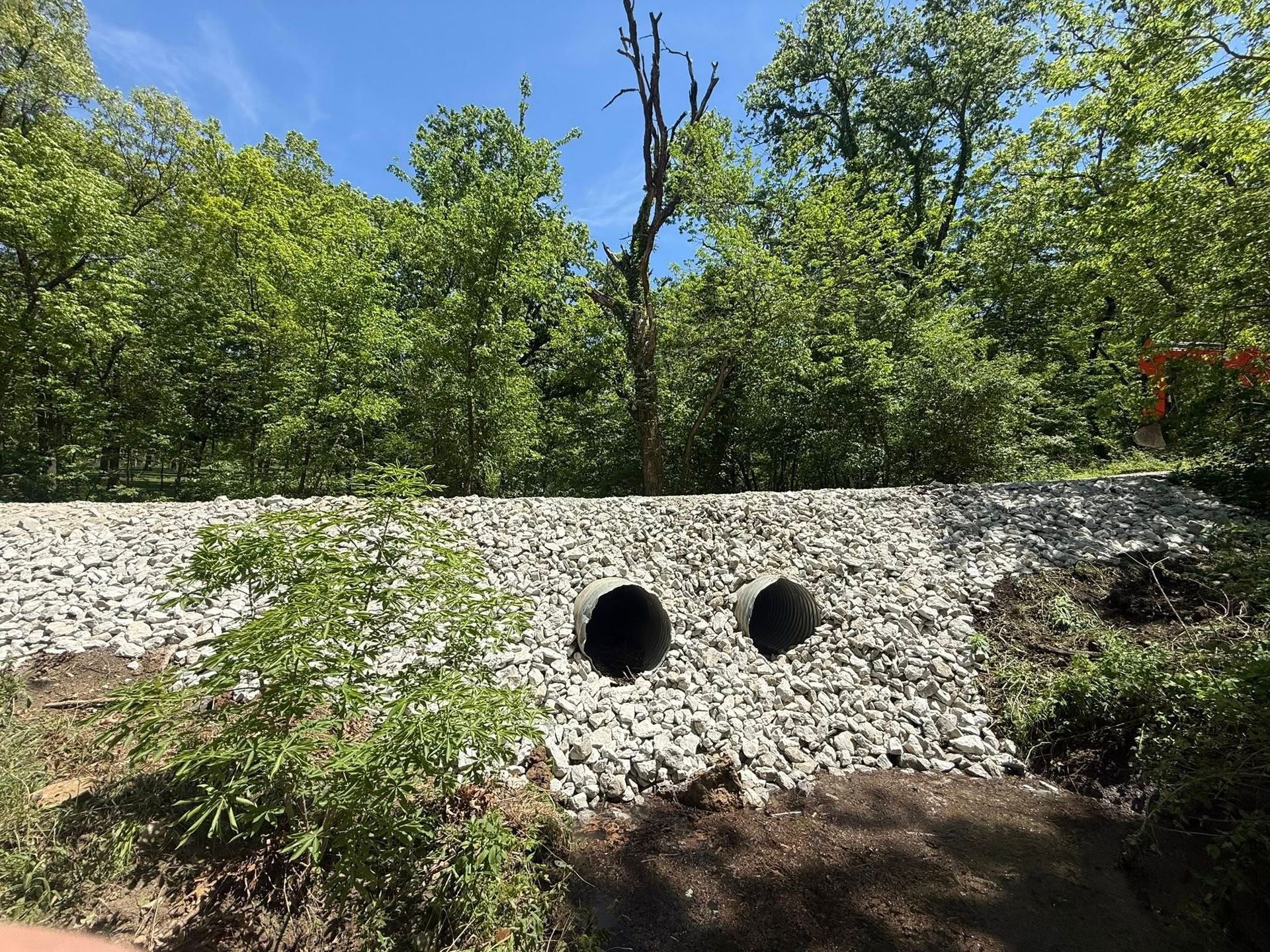 Concrete culvert wall with two round drainage pipes amid green trees and brush