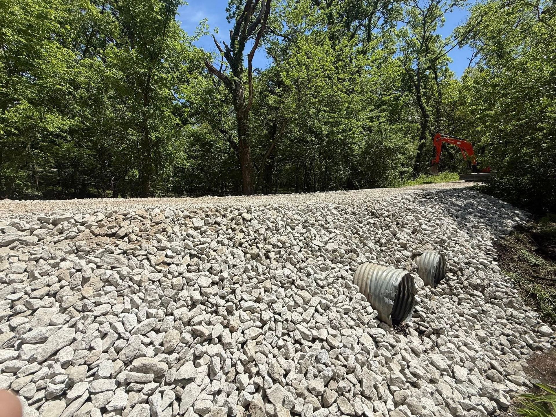 Rock-covered embankment with a drainage pipe, trees in the background, and a red flower at the top right