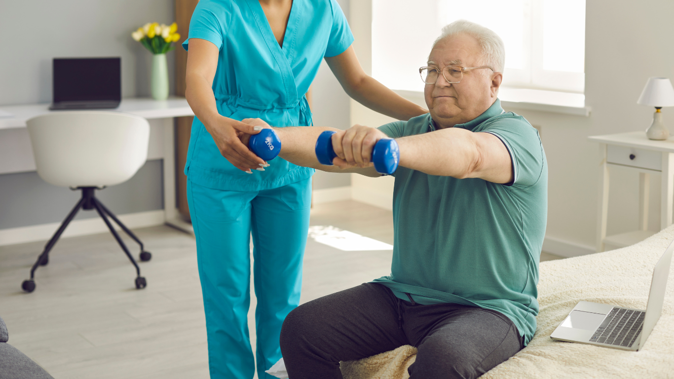 Man in blue scrubs explains medication to an older woman on a couch; they are in a home setting.