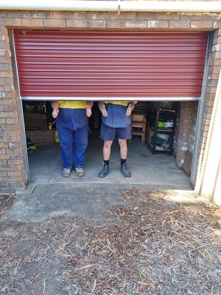 Two men are standing in a garage behind a red roller garage door.
