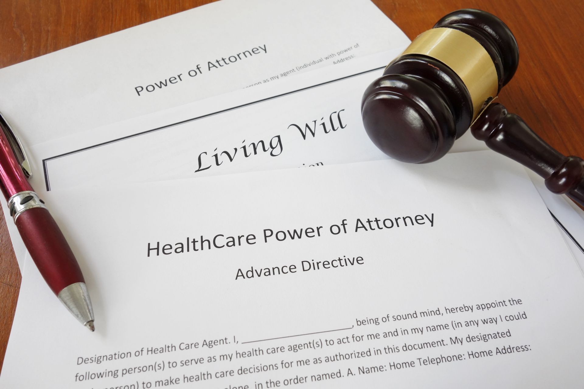 A gavel, a red pen, and legal forms for a Power of Attorney, Living Will, and Healthcare Power of Attorney on a desk. A gavel, a red pen, and legal forms for a Power of Attorney, Living Will, and Healthcare Power of Attorney on a desk.