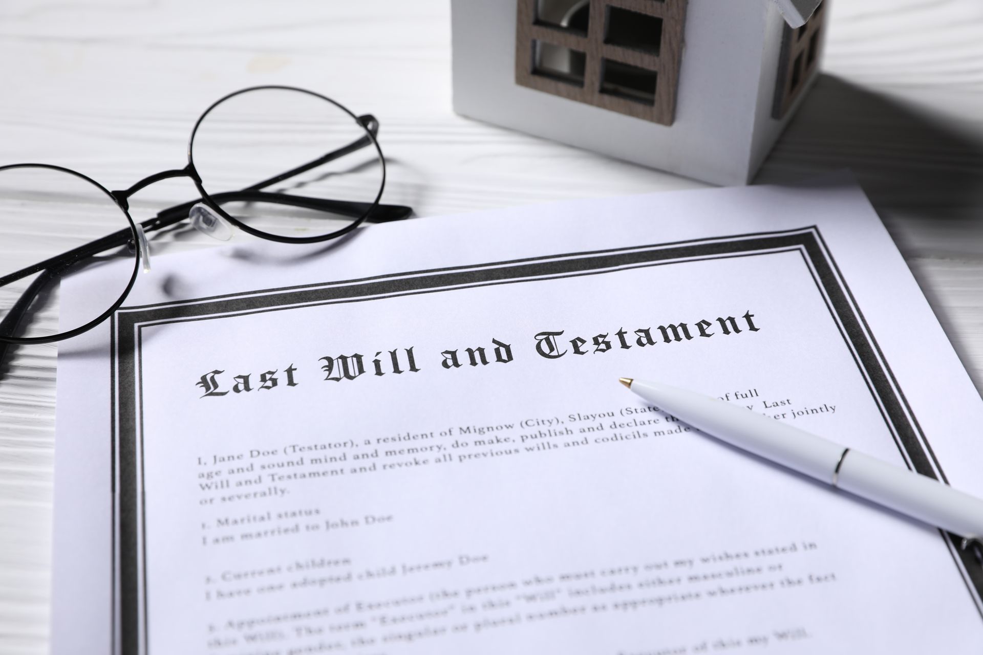 A Last Will and Testament document with a pen, eyeglasses, and a miniature house model on a white wooden surface.