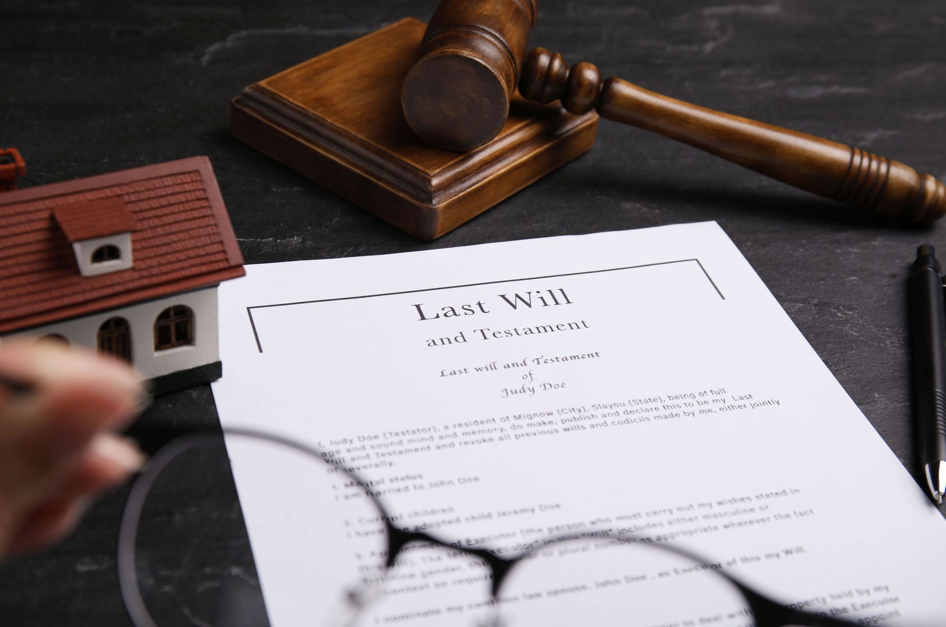 A Last Will and Testament document sits on a dark table next to a model house, a wooden gavel, and blurred eyeglasses. A Last Will and Testament document sits on a dark table next to a model house, a wooden gavel, and blurred eyeglasses.