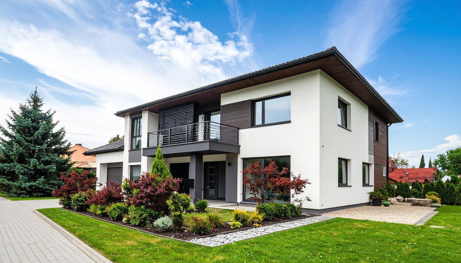 Modern two-story house with white exterior, dark trim, balcony, and landscaped yard under a blue sky.