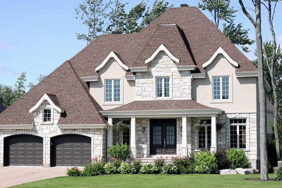 Two-story house with stone facade, brown roof, and arched garage doors; lush green lawn.