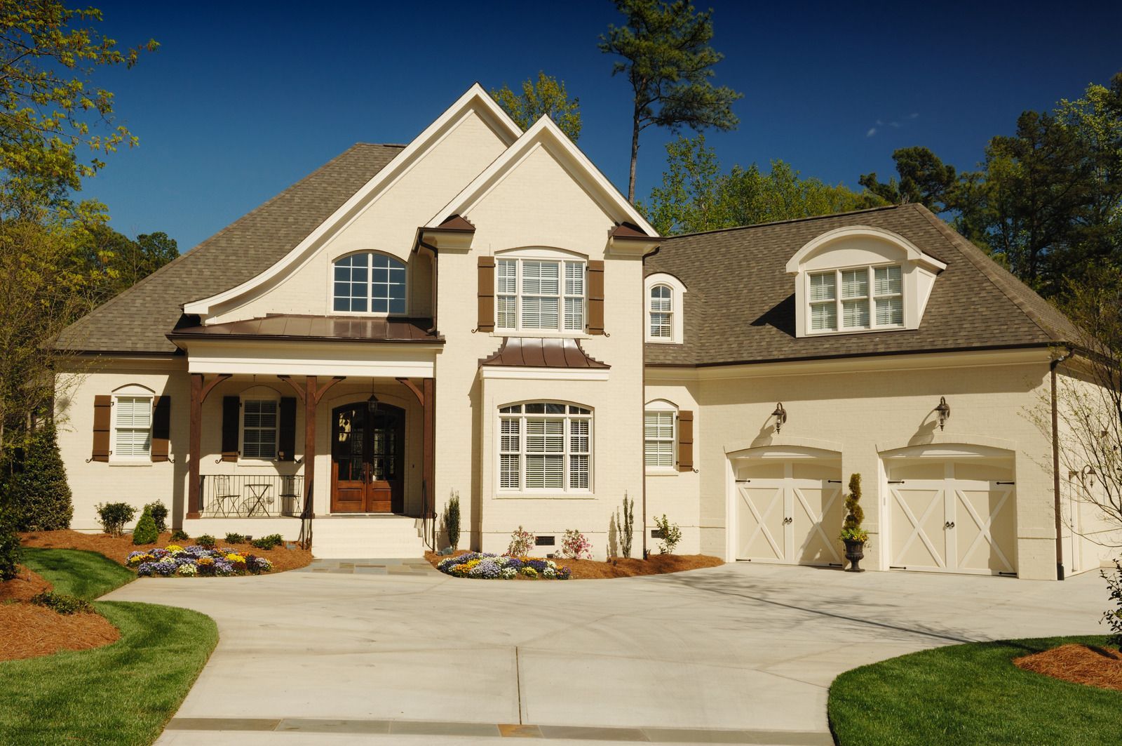 Beige two-story house with a driveway, brown shutters, and two garage doors on a sunny day.