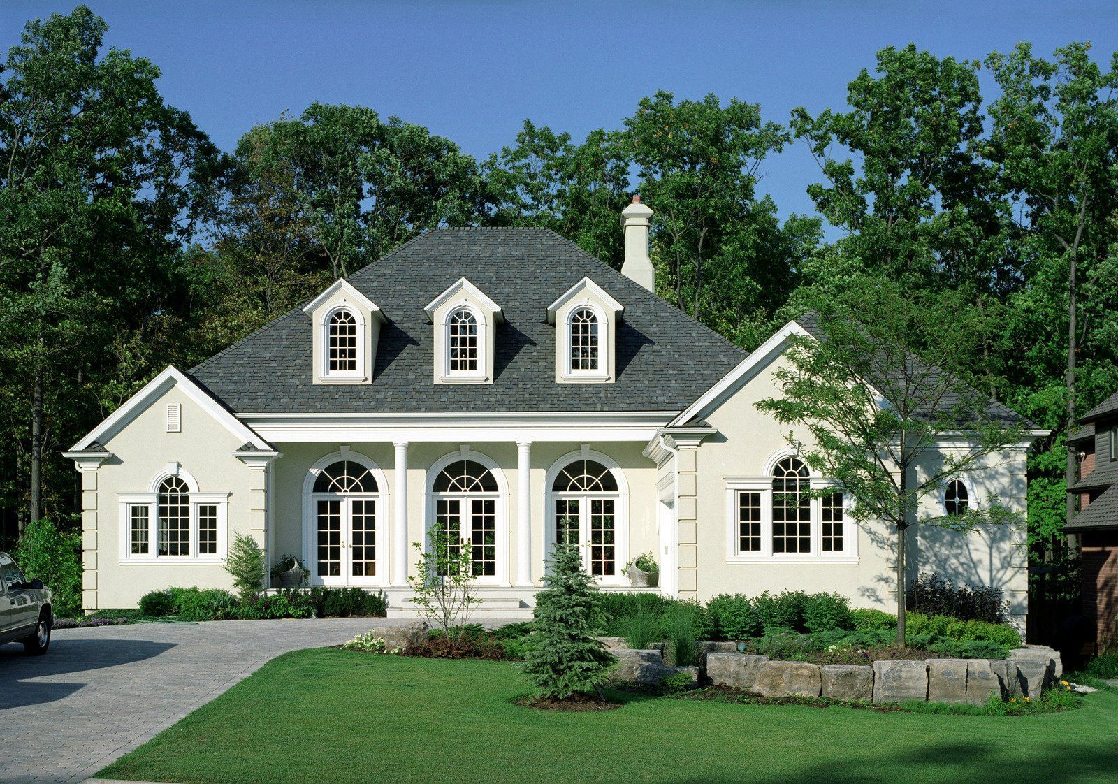 Beige house with gray roof, three dormer windows, and white trim, surrounded by trees and green lawn.