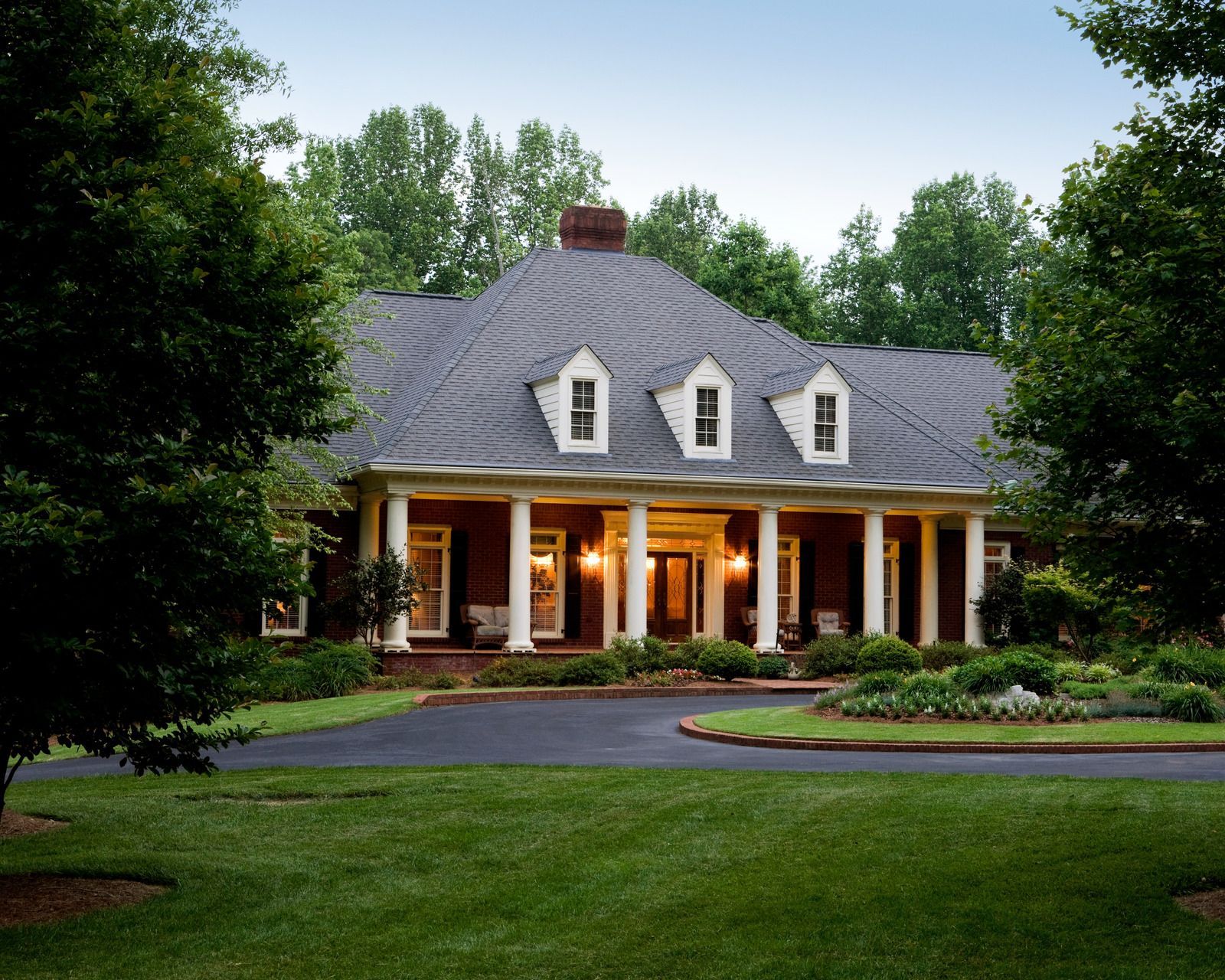 Large house with white columns, dormers, and a circular driveway on a grassy lawn, surrounded by trees.