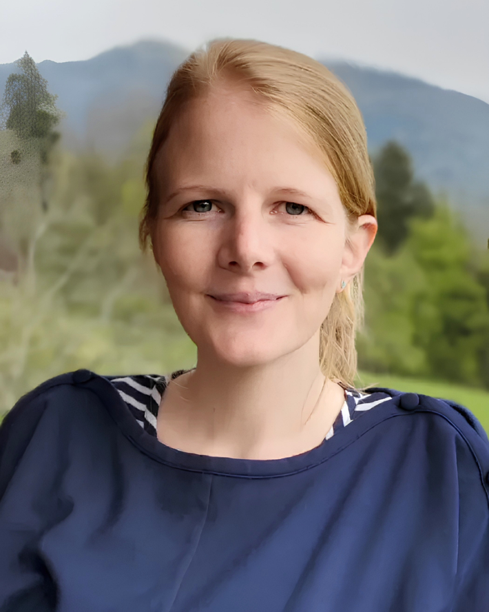 Personne souriante vêtue d'un haut bleu marine, avec une montagne et des arbres flous en arrière-plan.