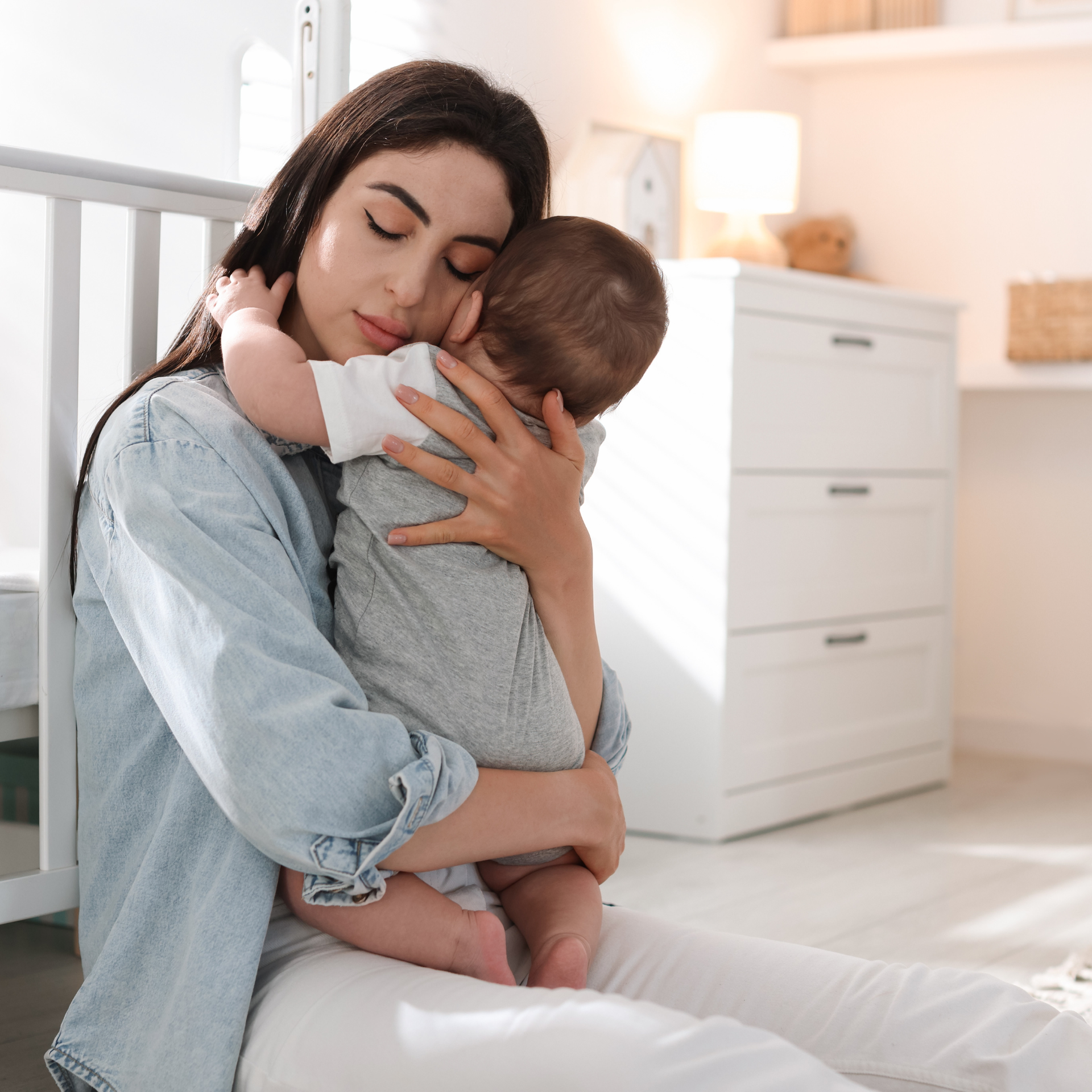 Une femme câline un bébé dans une chambre d'enfant lumineuse, à côté d'un berceau et d'une commode blancs.