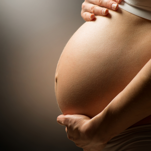 Gros plan sur un ventre de femme enceinte soutenu par des mains, sur un fond brun clair.