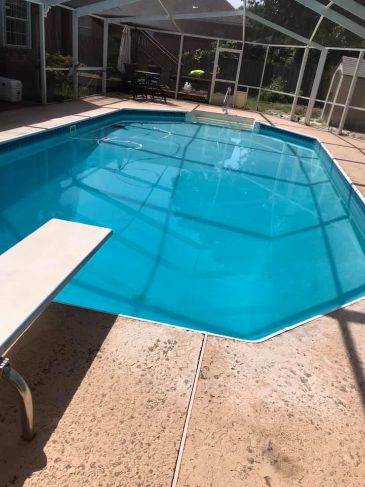 Swimming pool with diving board, screened enclosure; blue water, beige patio, sunny outdoor setting.