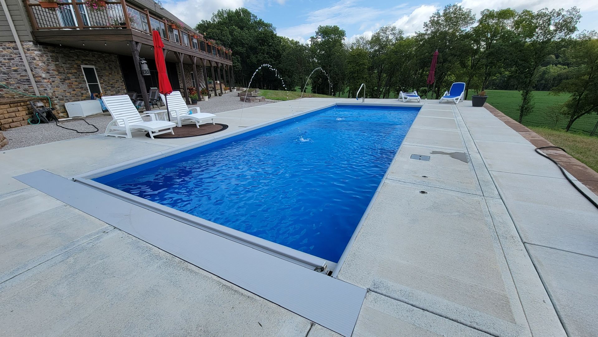 Rectangular blue swimming pool beside a patio and lounge chairs, surrounded by trees and lawn