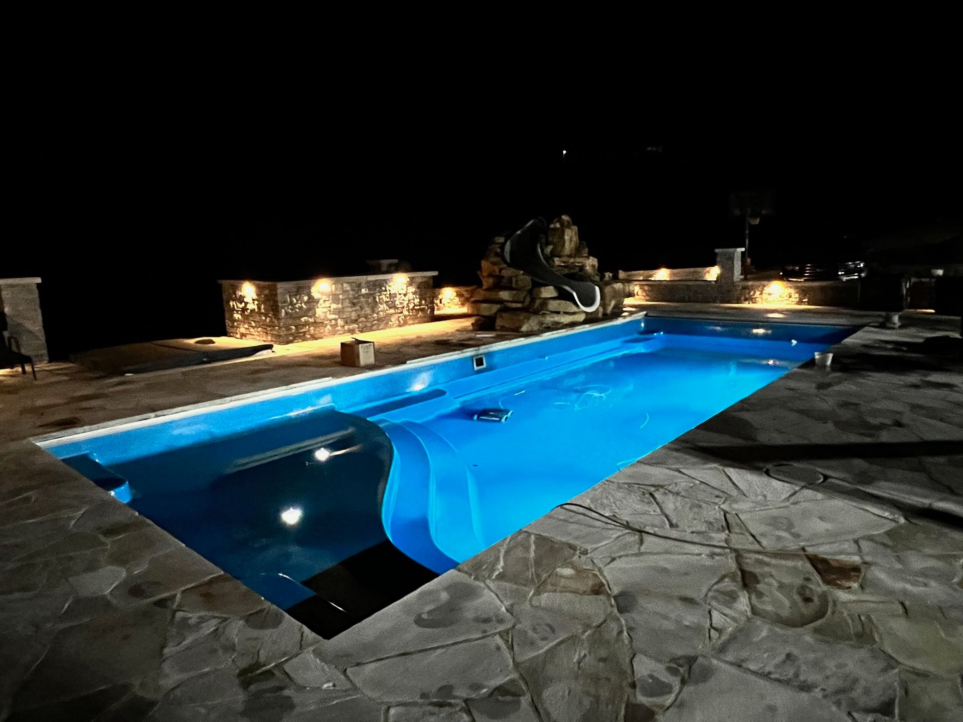 Nighttime outdoor pool with bright blue water, stone patio, and lit waterfall feature