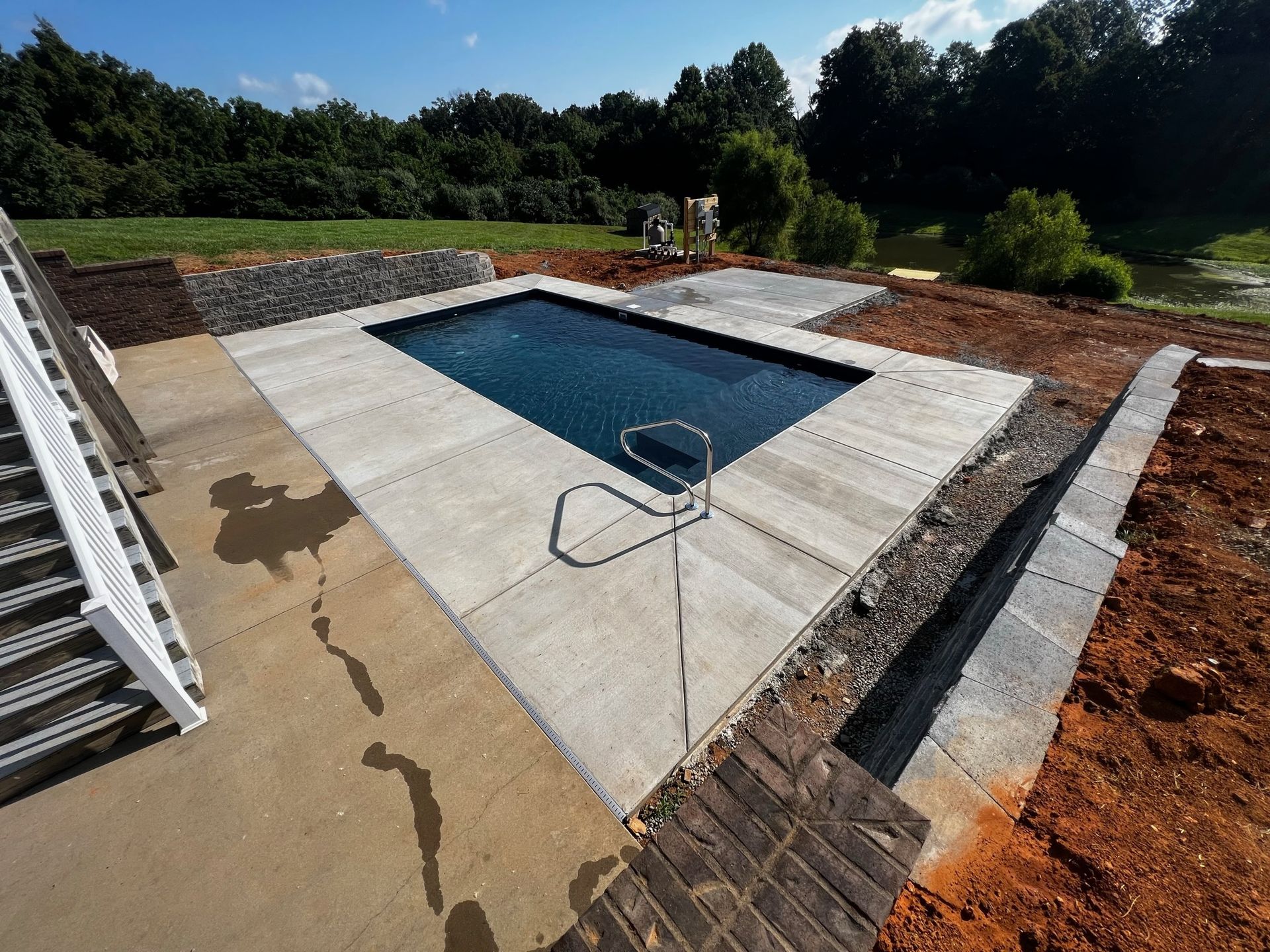 Rectangular swimming pool under construction, surrounded by concrete and dirt with trees in the background
