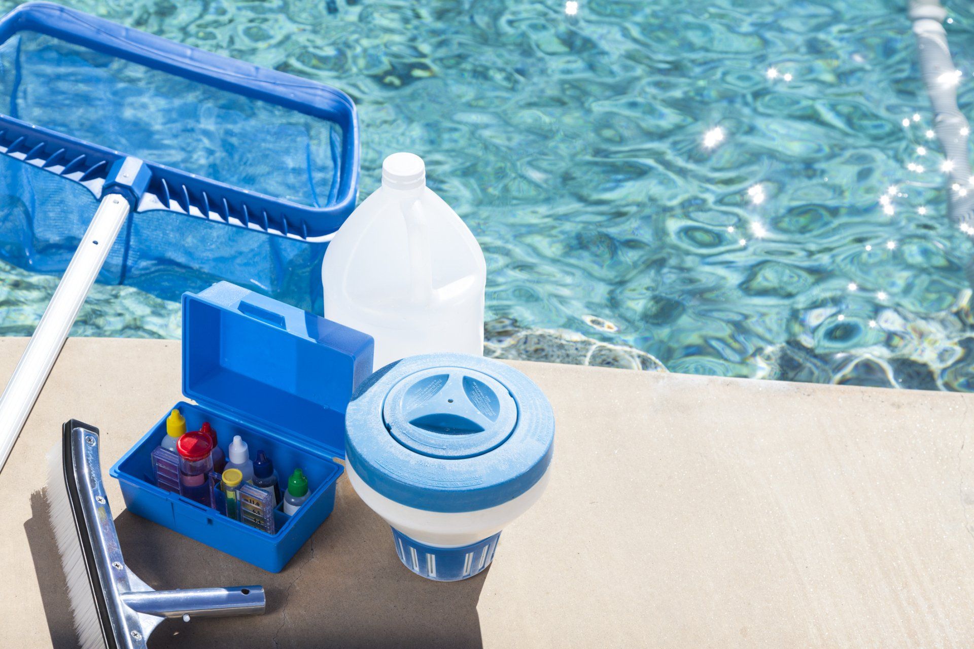 Pool cleaning supplies beside a swimming pool, including a net, brush, bottle, and chlorine float