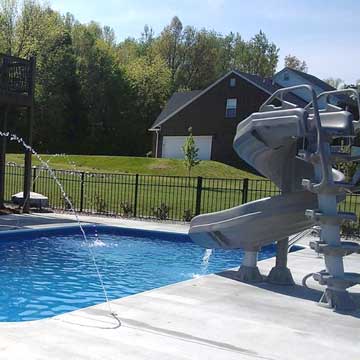Backyard pool with a water slide, patio deck, and green lawn under a sunny sky
