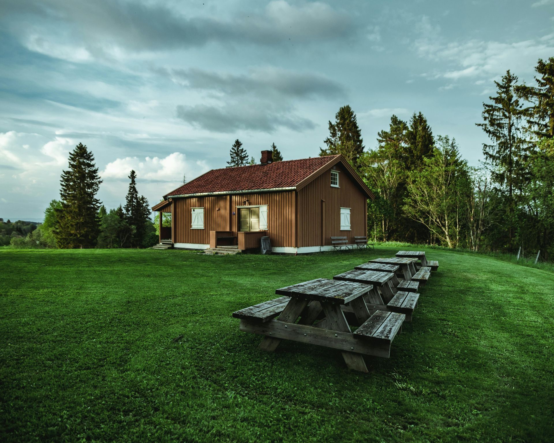 Wooden cabin with picnic tables on green grass under a cloudy sky.