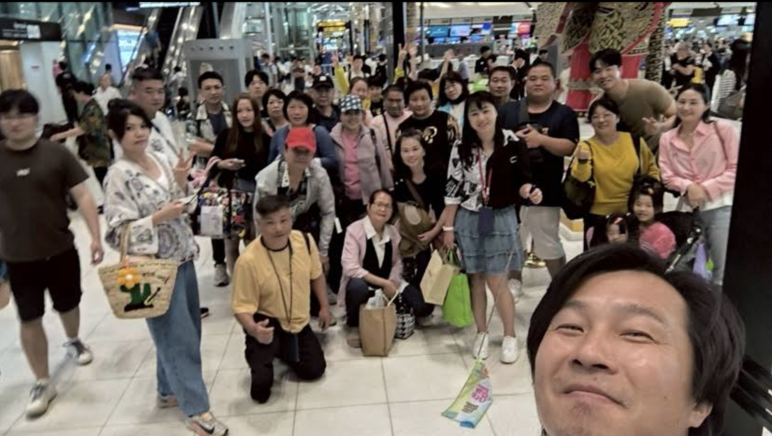 在明亮的室內商場裡，一群人擺姿勢自拍，笑容滿面，購物袋清晰可見。