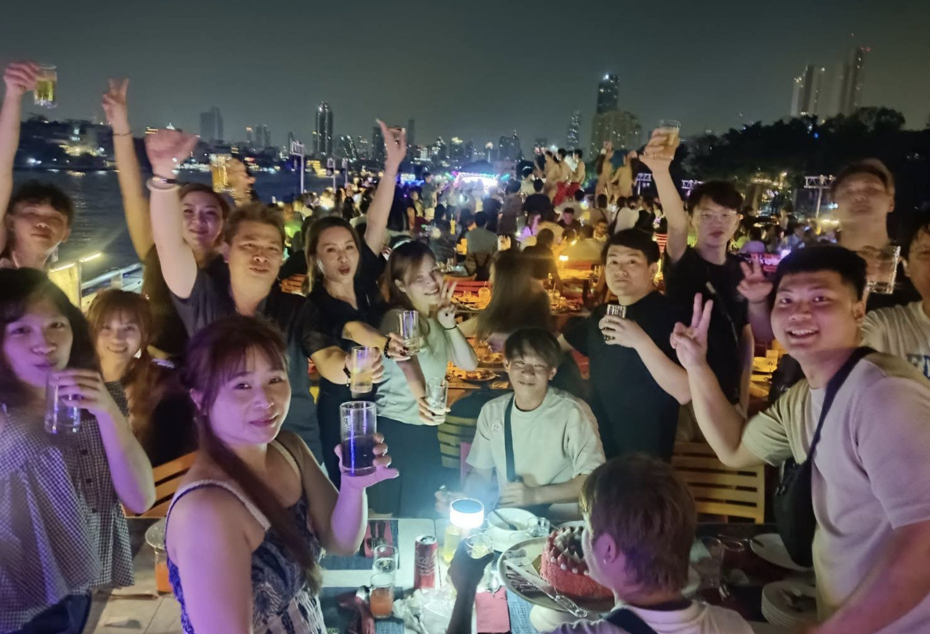 一群人在夜間屋頂聚會上擺姿勢拍照，他們面帶微笑，舉起和平手勢，身後是璀璨的城市燈光。