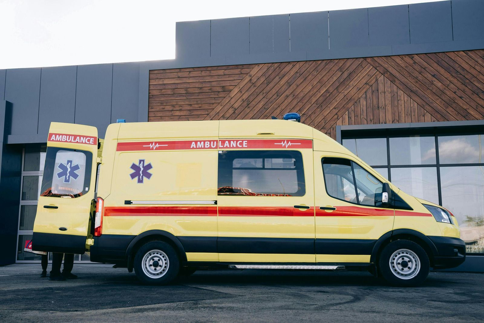 Yellow ambulance with red stripes and star of life symbol, doors open, parked in front of a building.
