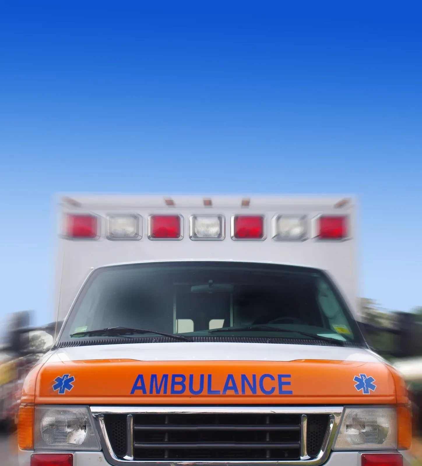 Ambulance speeding, orange and white vehicle with flashing lights, against a blue sky.