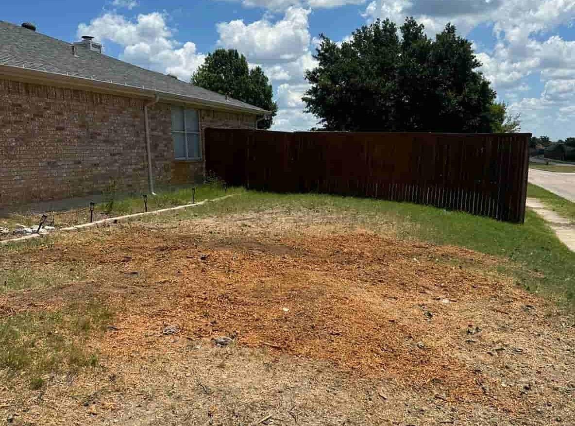 Bare patch of dirt in a yard next to a brick house and wooden fence under a blue sky.