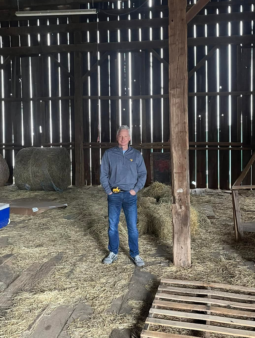 Person standing in a barn with a large tree trunk and a hay-covered floor.