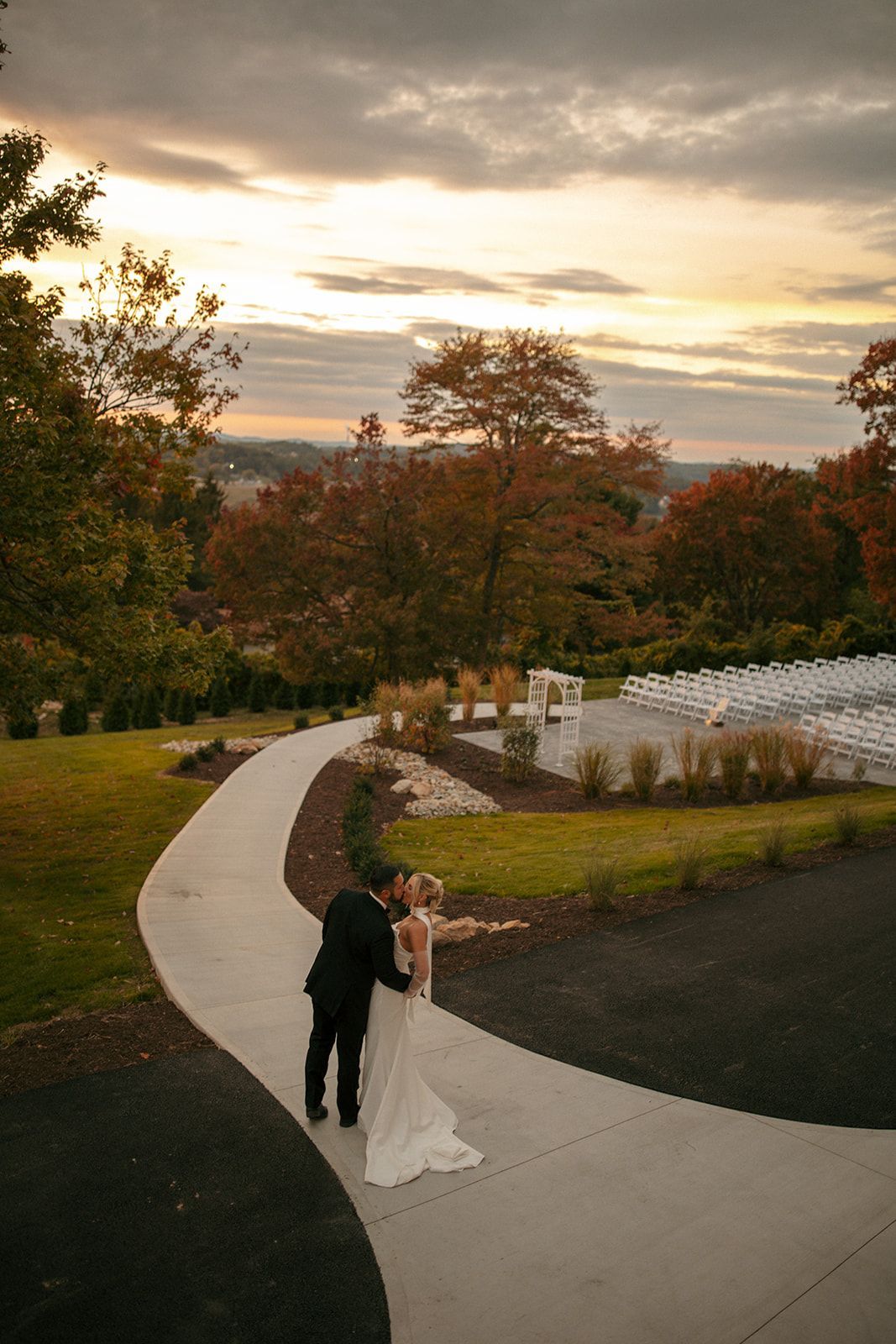 Couple in wedding attire on a winding path at sunset in a park with autumn trees and water nearby