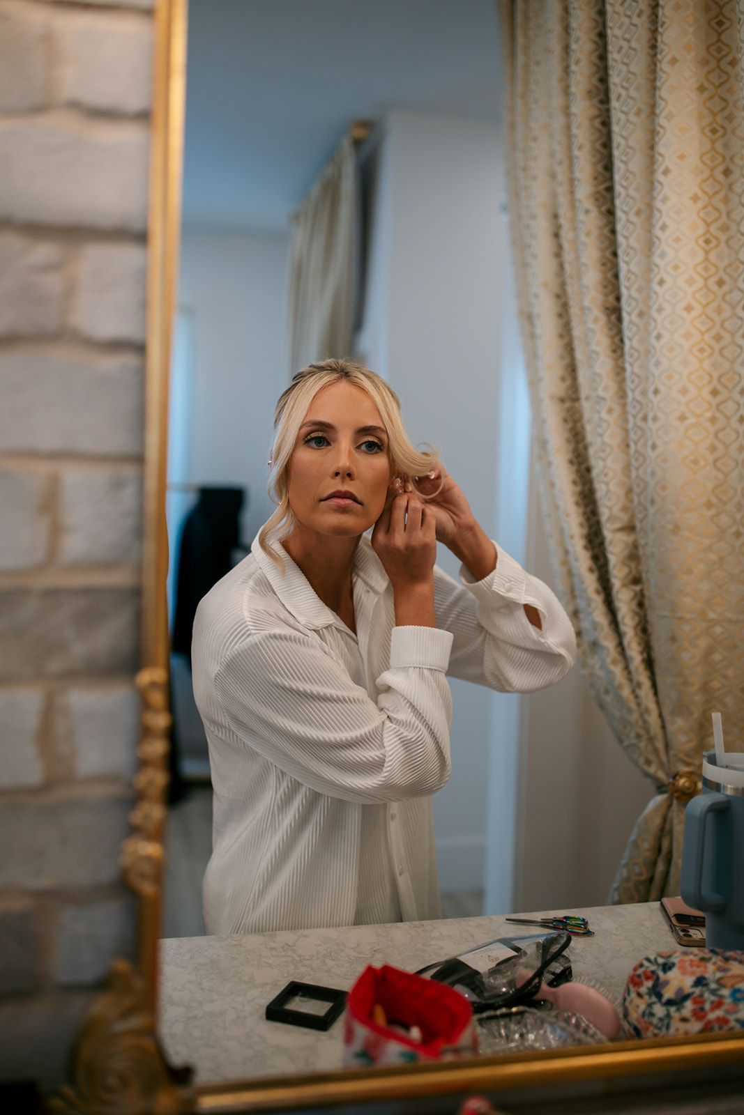 Woman in a white robe adjusting earrings at a vanity mirror, with makeup items and a curtain nearby.