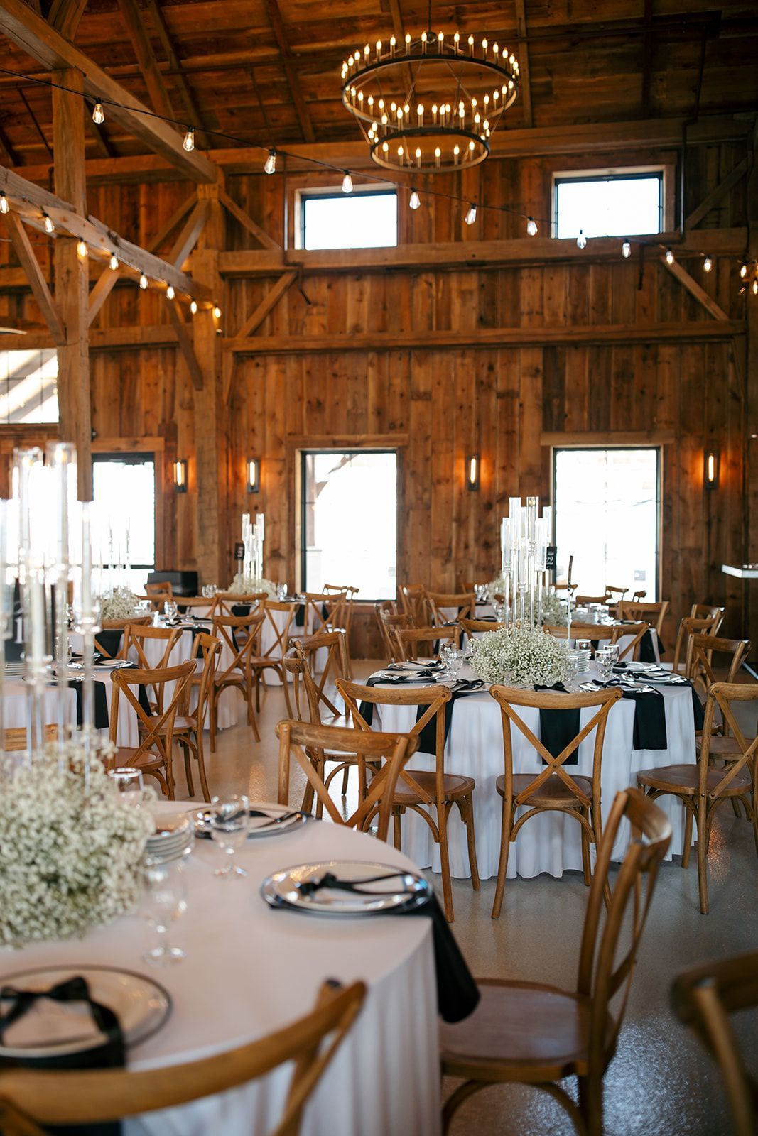 Rustic barn-style banquet hall with wooden tables, chairs, and white floral centerpieces under chandeliers