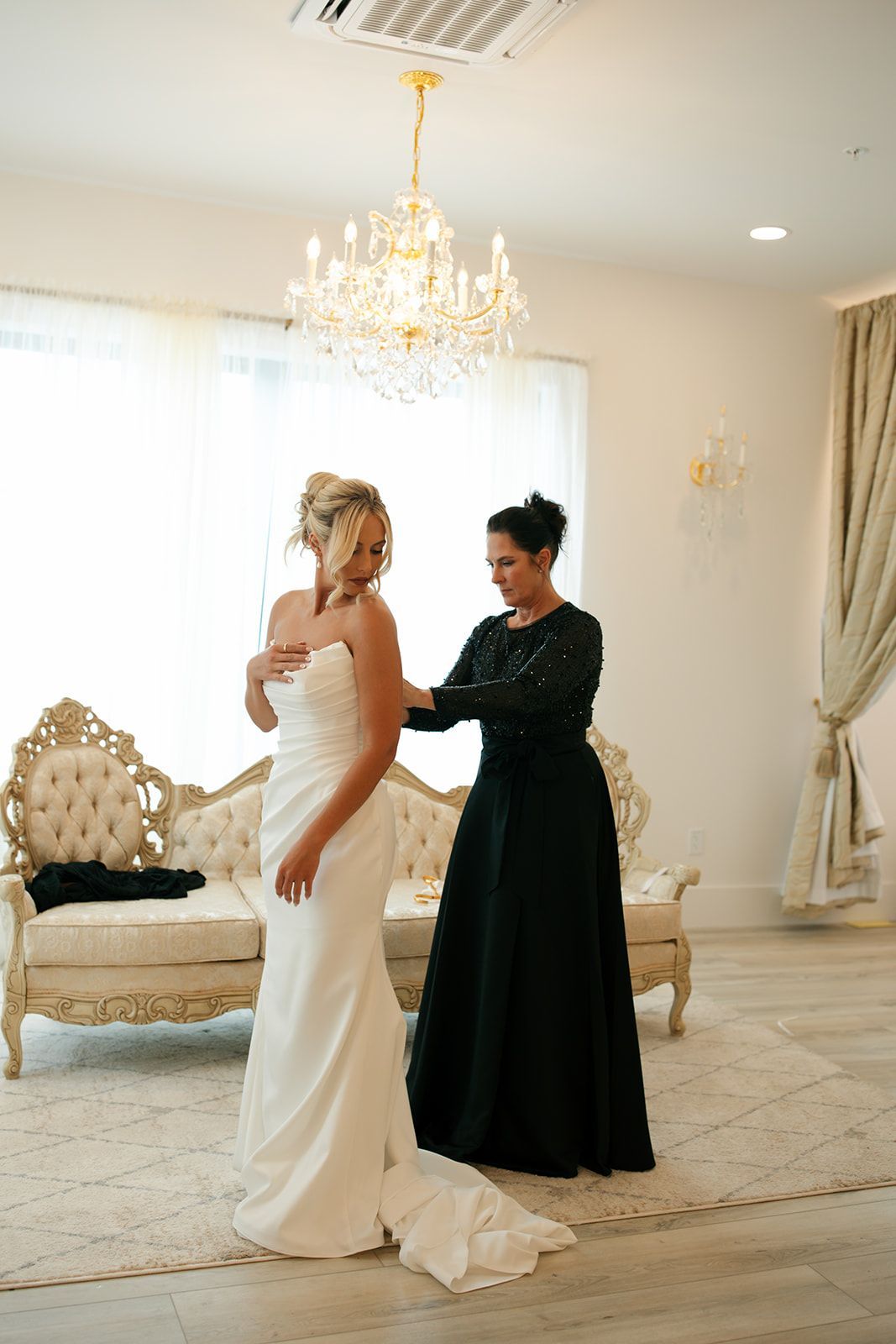 Two women in formal gowns in a bright ballroom, one adjusting the other’s dress train.