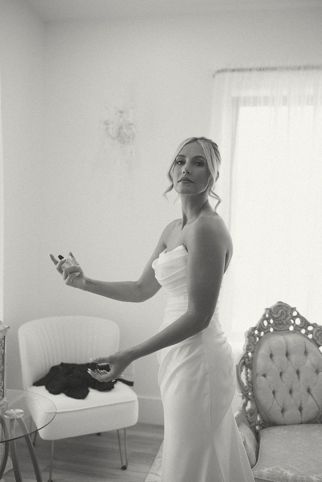 Person in a white dress standing in a living room, holding a small object near a chair.