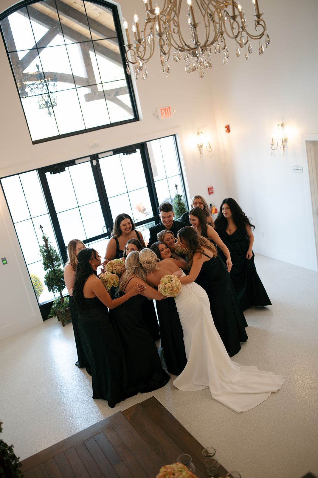 Bridal party in black dresses surrounds a bride in a white gown inside a bright, elegant room.