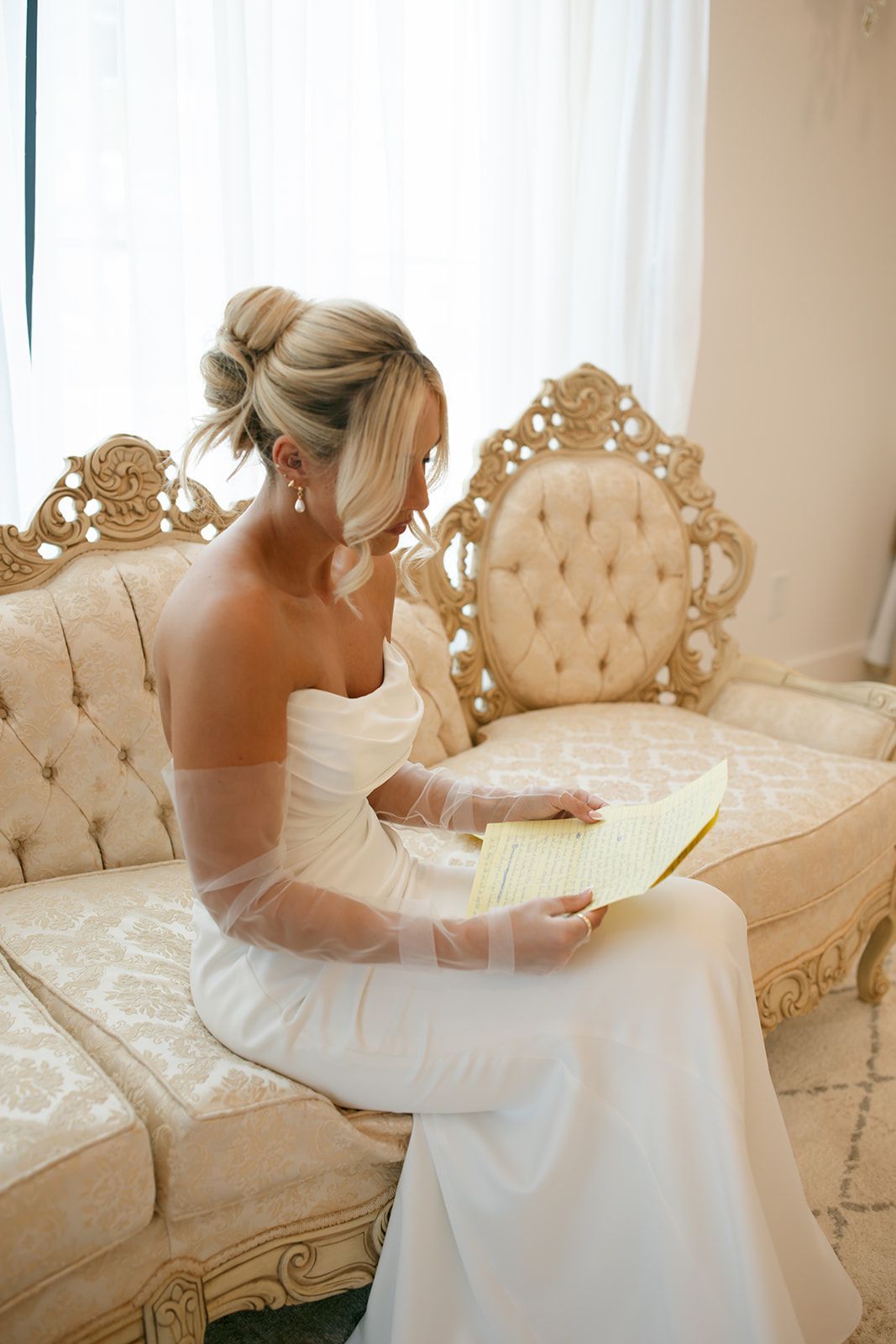 Bride in a white gown seated on an ornate sofa, holding a bouquet in a softly lit room