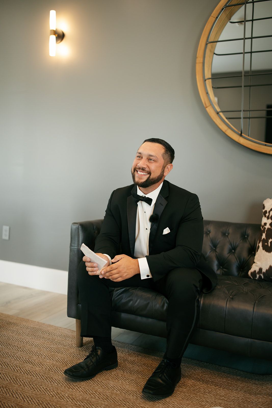 Man in a black suit sits smiling on a black couch in a modern room with a wall sconce and round mirror.