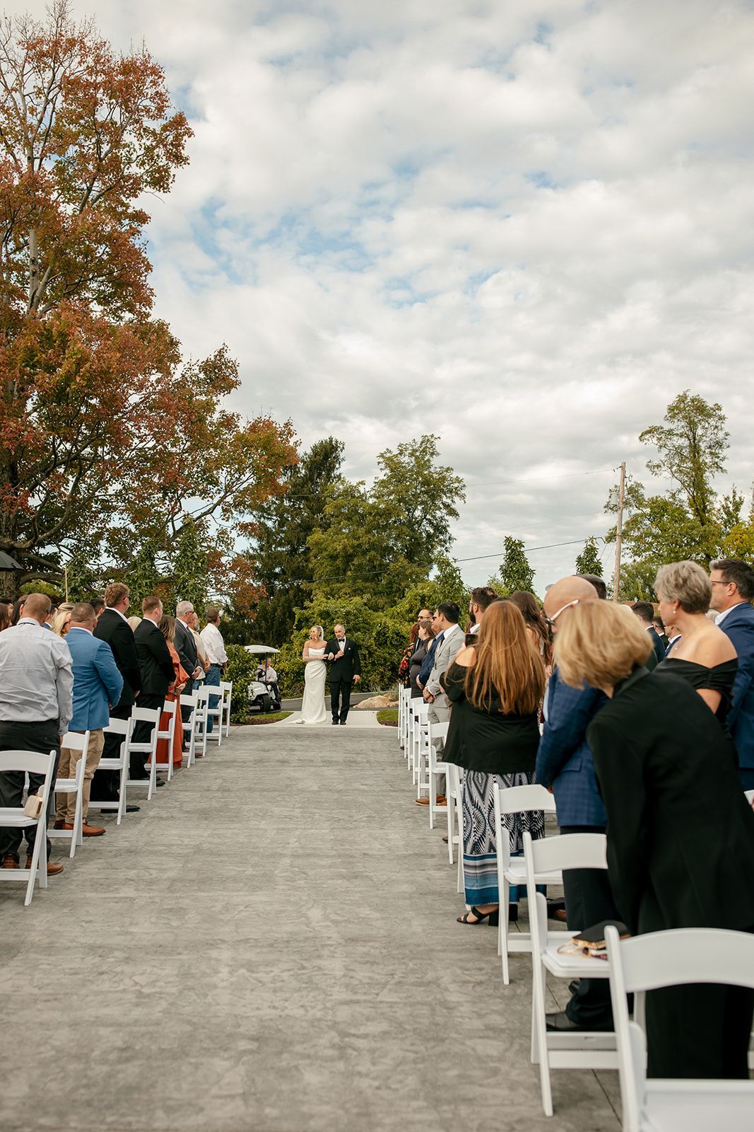 Guests seated outdoors on white chairs, facing a ceremony aisle under autumn trees and cloudy sky